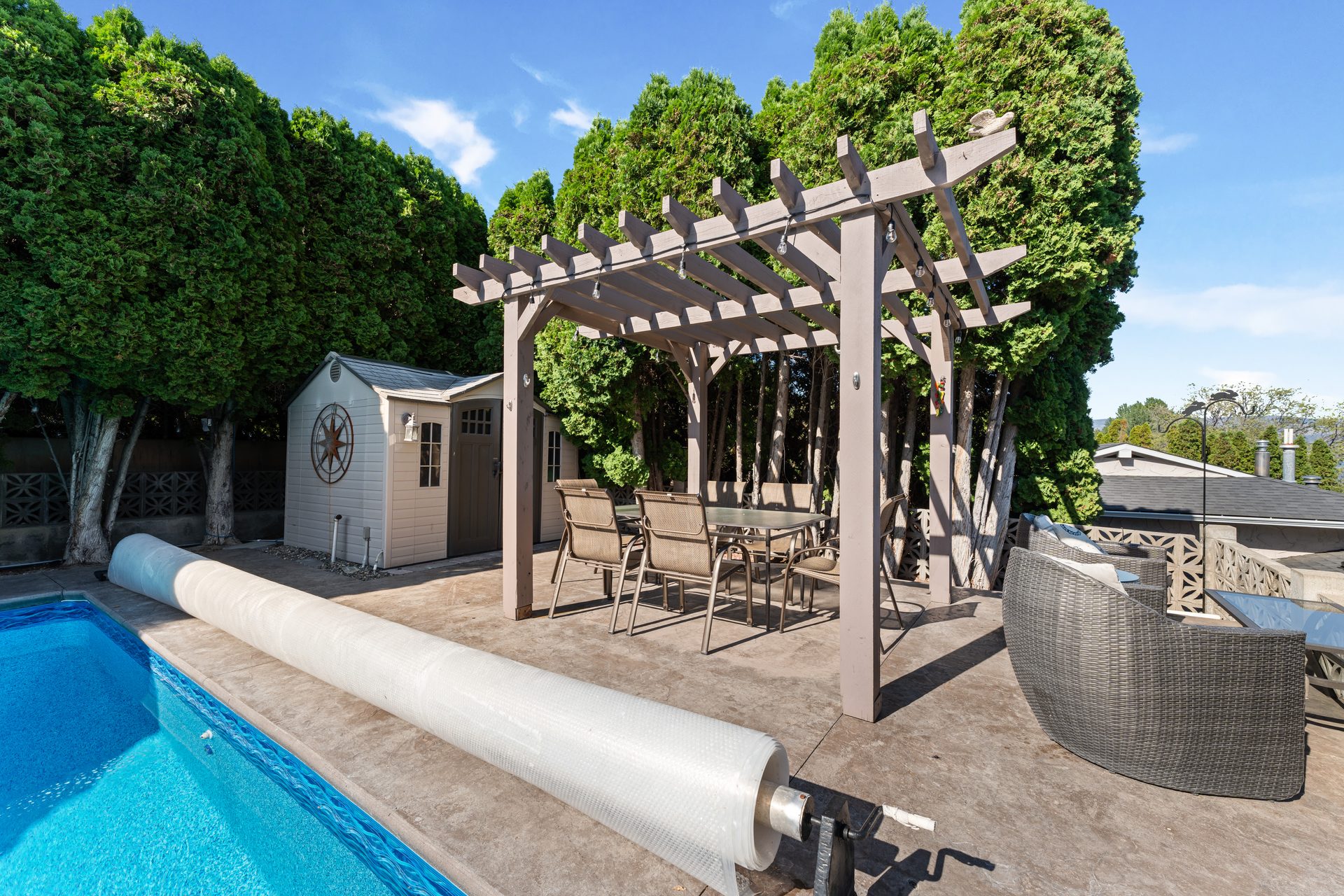 Pergola dining area with string lights beside pool, shed and cedar hedges