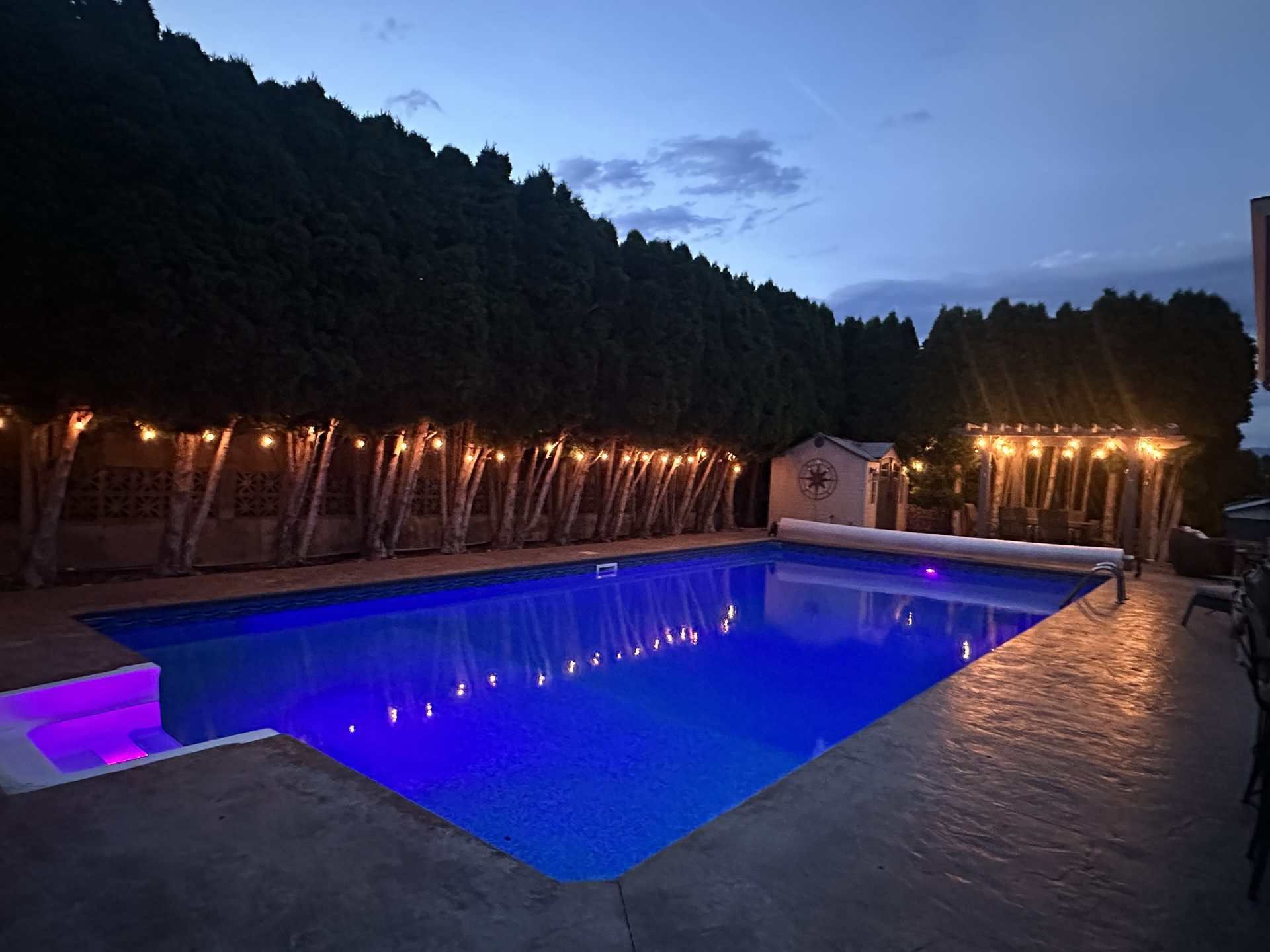 Private pool lit up at night with colour-changing lights and string lights over the patio